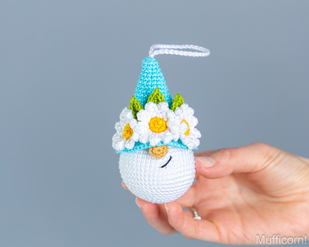 A hand holds a small crocheted white snowman ornament wearing a light blue pointed hat decorated with white daisies that have yellow centers and green leaves. The background is a solid, neutral gray.