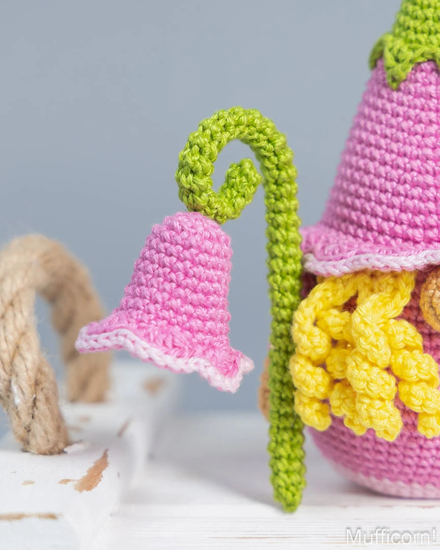 A close-up of a crocheted gnome figure with a pink hat, yellow beard, and green stem holding a pink bell, set against a soft gray background with a white wooden surface.