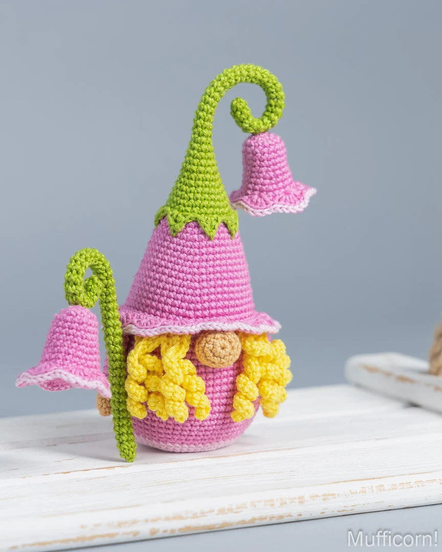 A crocheted gnome doll with a pink hat and bell, yellow curly hair, and green pointed hat, displayed on a white wooden surface against a soft gray background.