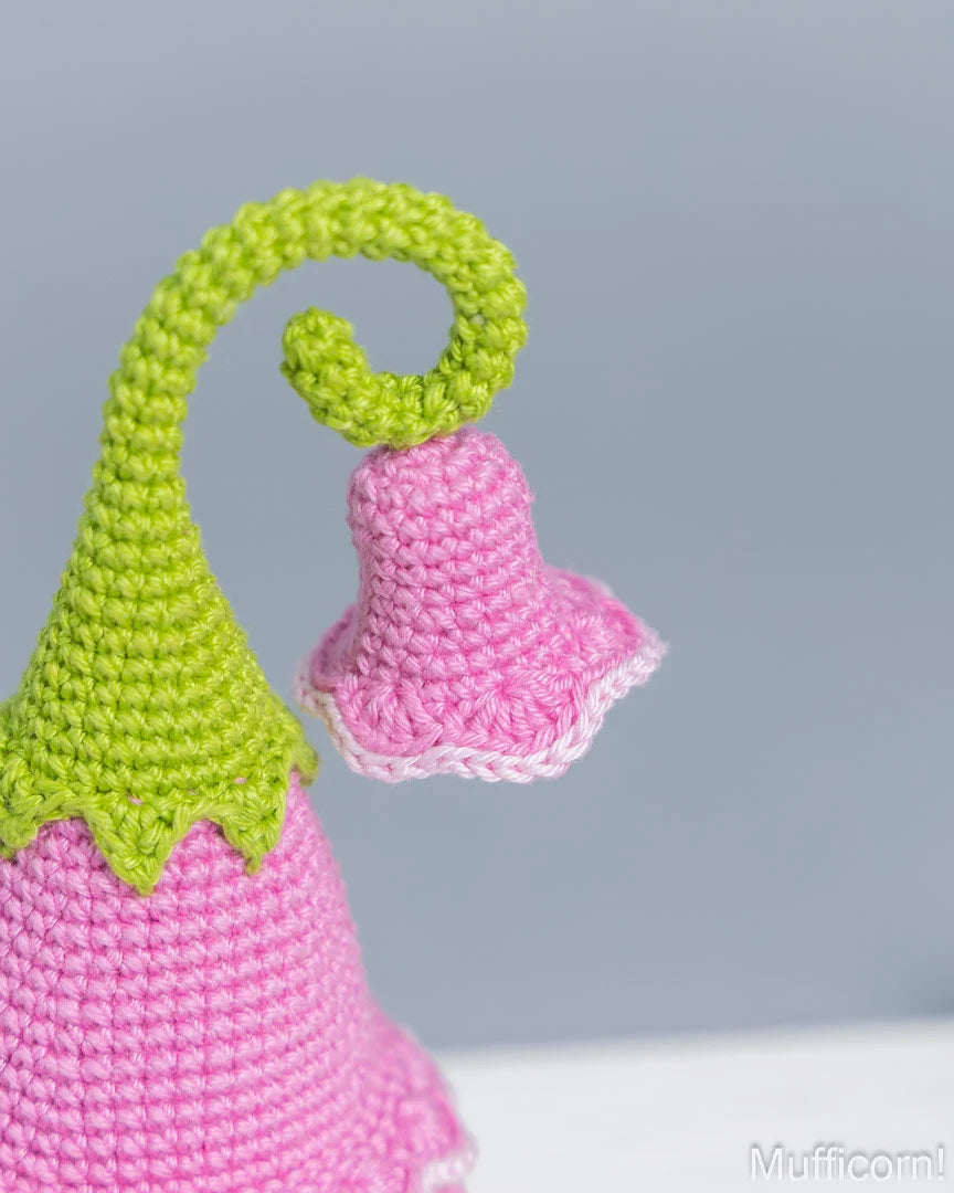 Close-up of a crocheted flower decoration with a pink body and a curled bright green stem, photographed against a soft light gray background.