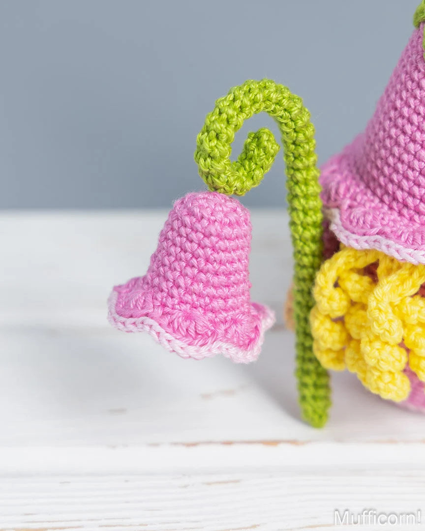 A close-up of a crocheted bell-shaped flower with a pink bloom, green stem, and yellow petals, set on a white wooden surface against a soft gray background.