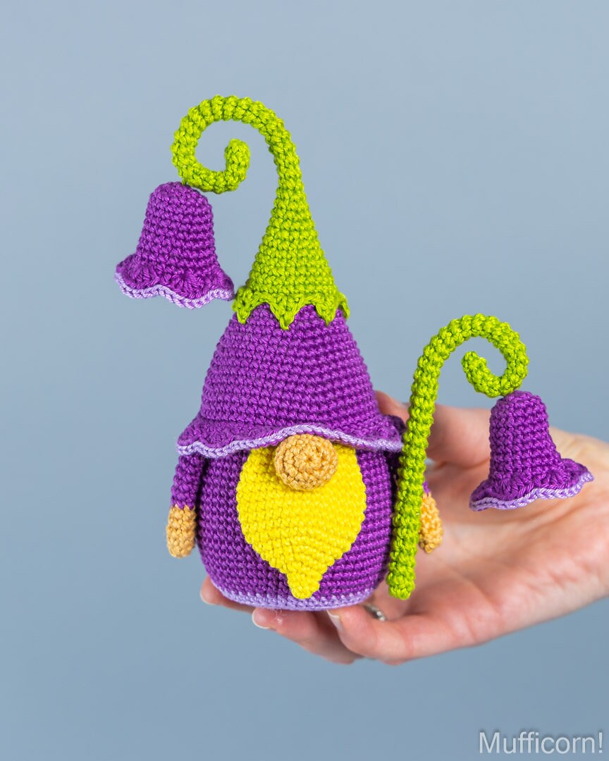A crocheted gnome doll with a purple body and hat, bright green pointed hat and curved stems, and a yellow beard, being held in a person's hand against a soft blue-gray background.