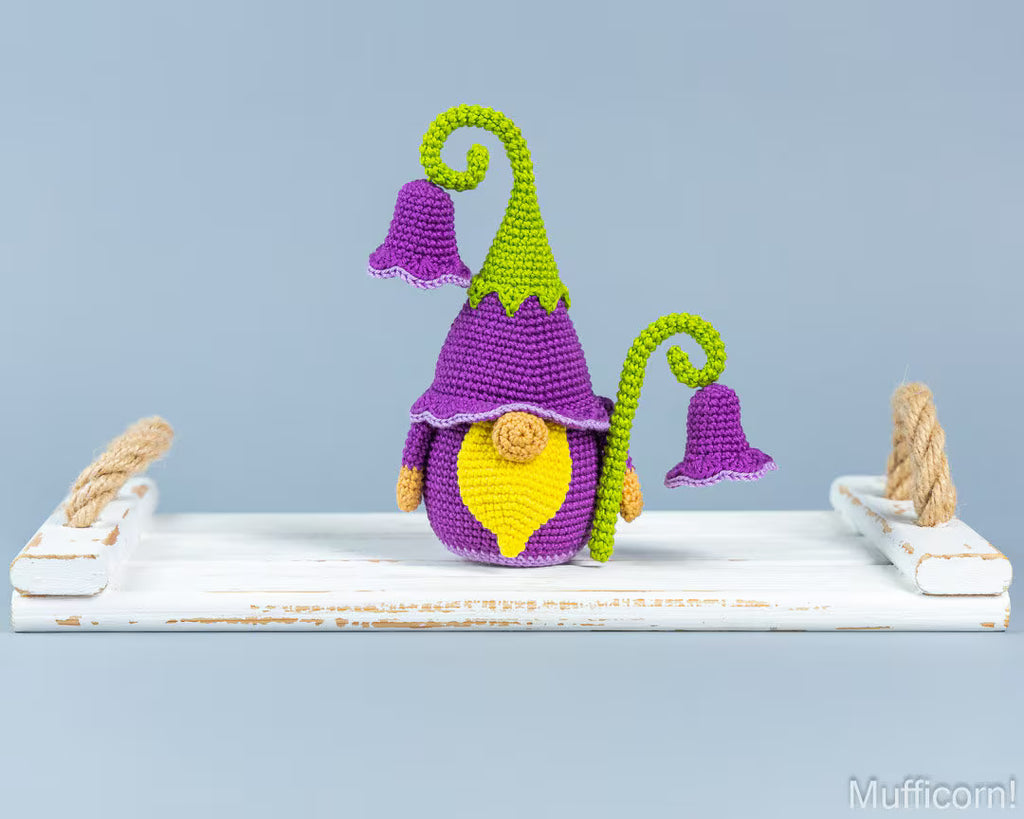 A crocheted gnome figurine with a purple hat and bell, green pointed hat and vine, and a yellow face, displayed on a white rustic wooden shelf against a light blue background.