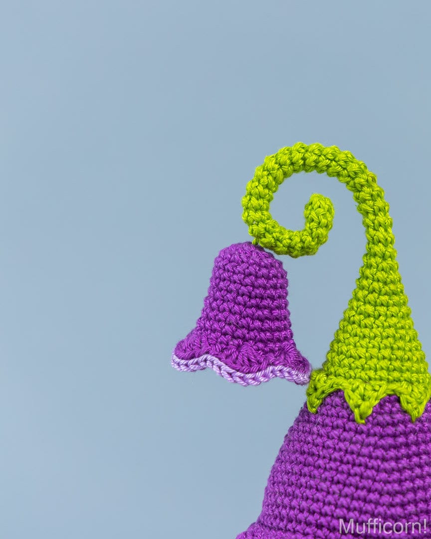 A close-up of a crocheted bell-shaped campanula in purple with a bright lime green curling stem, set against a solid light blue background.