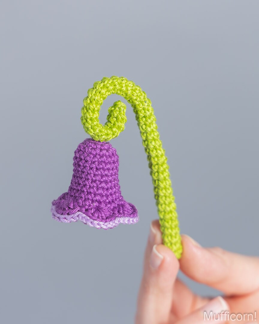 A small crocheted bell-shaped flower with a bright lime green stem and curling top, held by a person's hand against a plain light gray background.