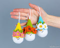 Three small crocheted gnome ornaments are held in a person's hand against a plain gray background. Each ornament has a white round body, a colorful pointed hat, and a flower crown.