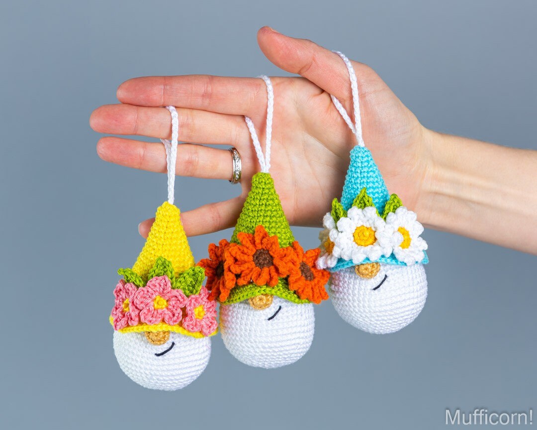 Three small crocheted gnome ornaments are held in a person's hand against a plain gray background. Each ornament has a white round body, a colorful pointed hat, and a flower crown.