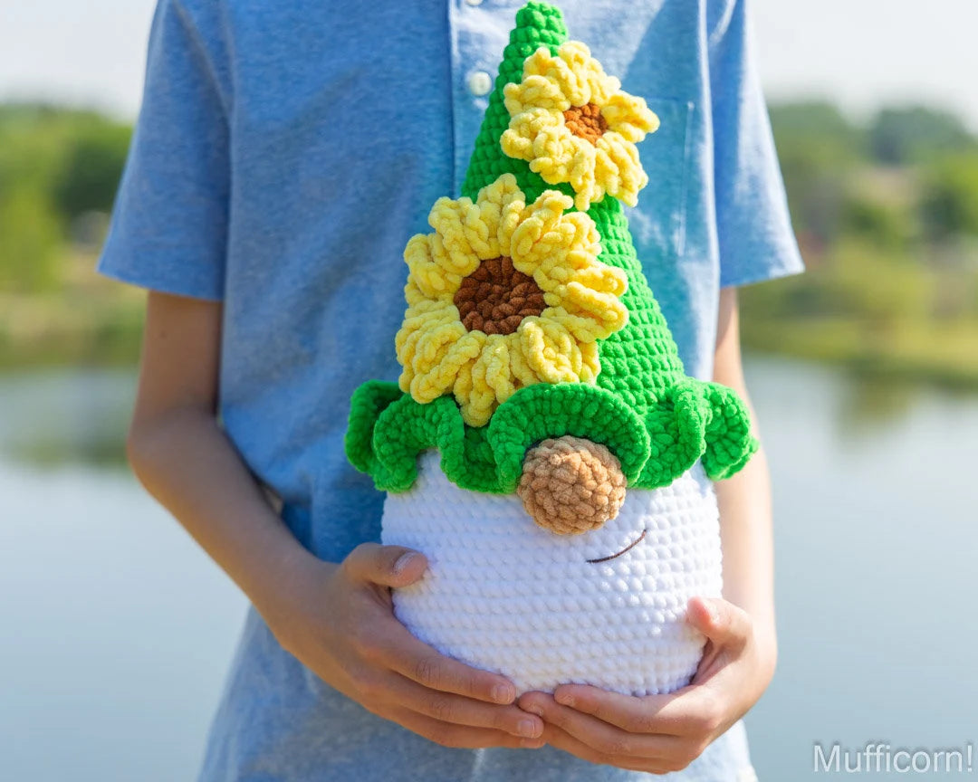 A person in a light blue shirt holds a crocheted sunflower gnome against a blurred outdoor background of water and trees. The gnome has a white body, a green pointed hat with yellow sunflowers, and a brown pinecone nose.