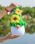 A crocheted plush gnome with yellow sunflowers and a green pointed hat, held up by two hands against a blurred outdoor background of water and trees.