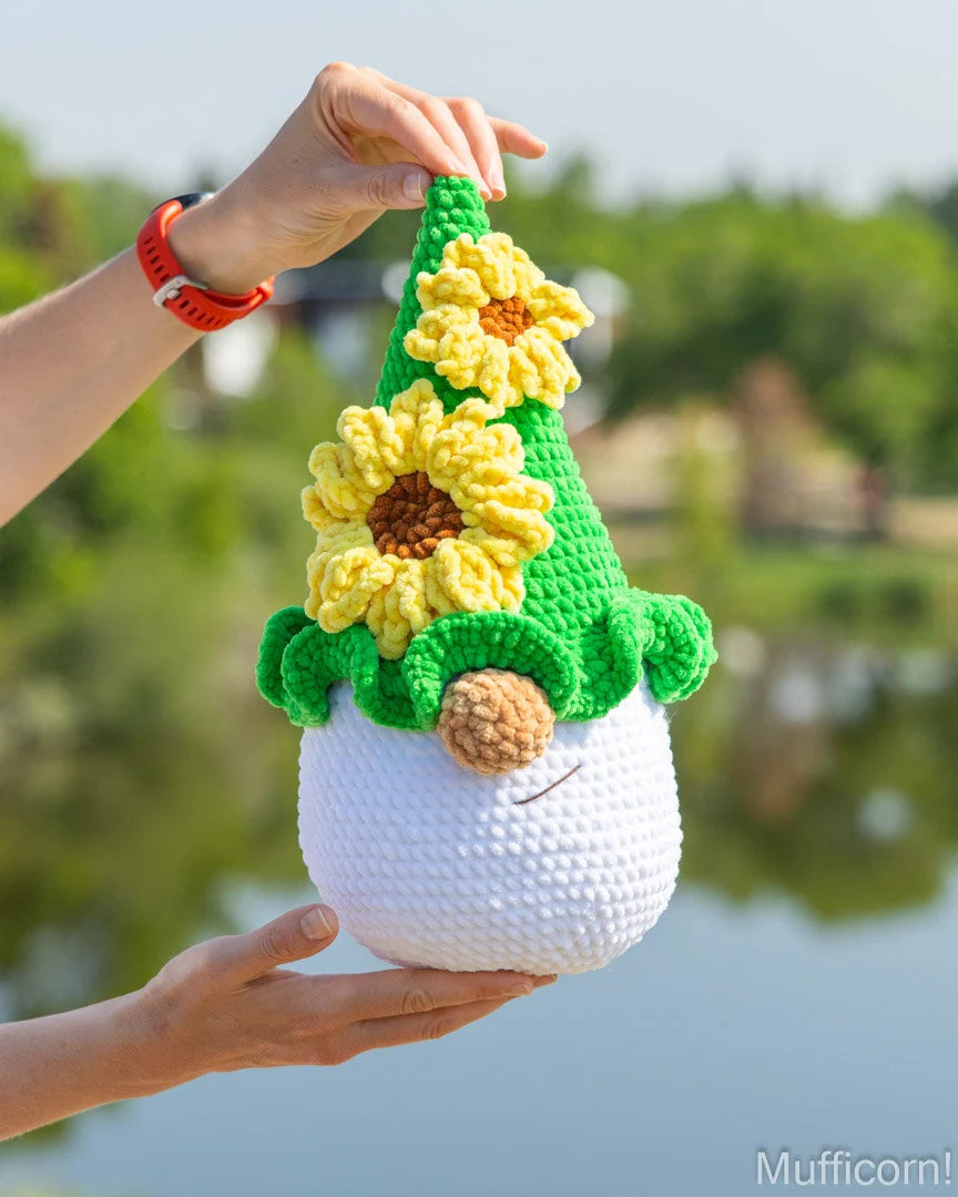 A crocheted plush gnome with yellow sunflowers and a green pointed hat, held up by two hands against a blurred outdoor background of water and trees.