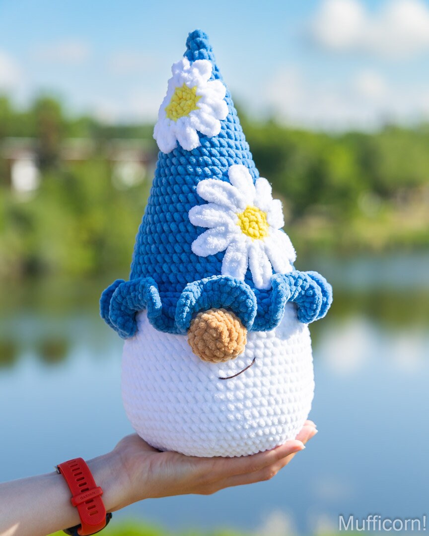 A crocheted plusgnome doll with a blue pointed hat, white daisy flowers, and a brown carrot nose is being held in a person's hand outdoors with a blurred lake and trees in the background. 