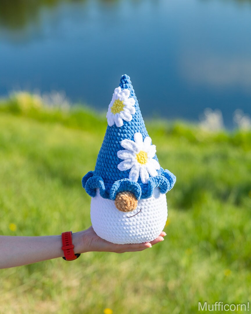 A crocheted gnome doll with a blue pointed hat decorated with white daisy flowers and a brown nose, held in a person's hand against a blurred outdoor background of green grass and blue water.