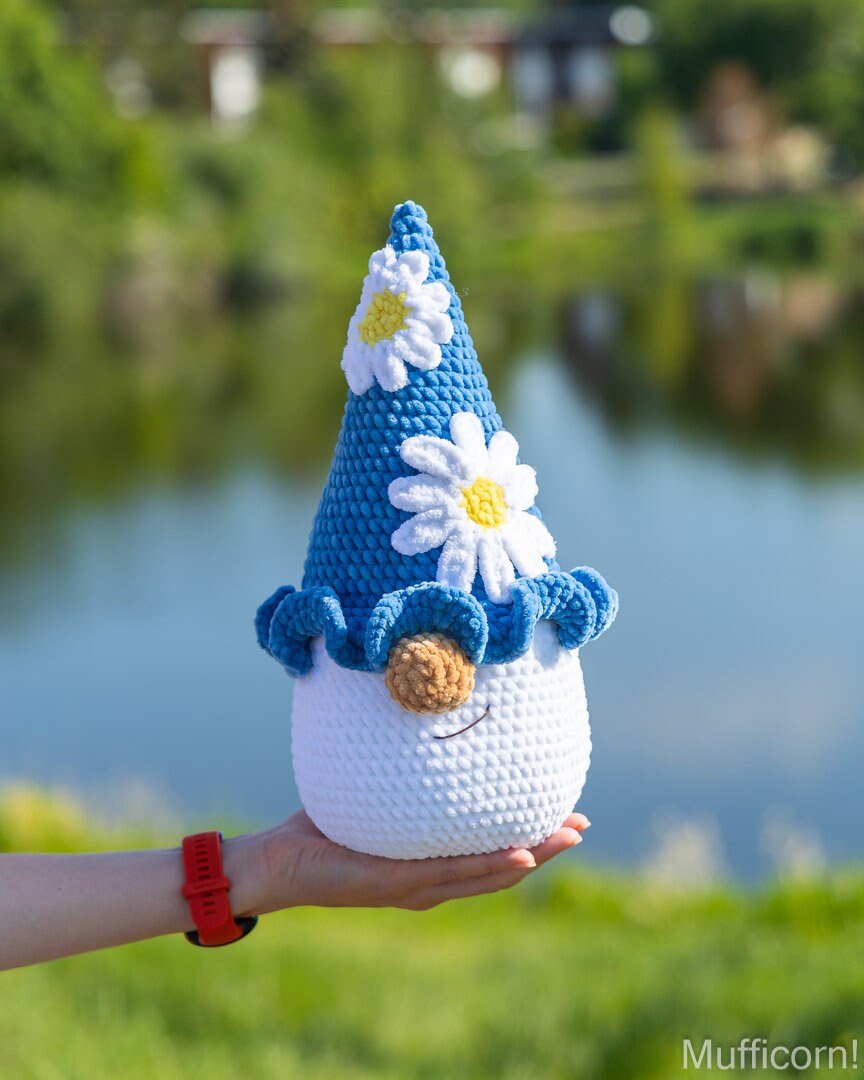 A crocheted plush gnome with a blue hat, white body, and daisy flowers is held in a person's hand outdoors in front of a blurred lake and greenery background.