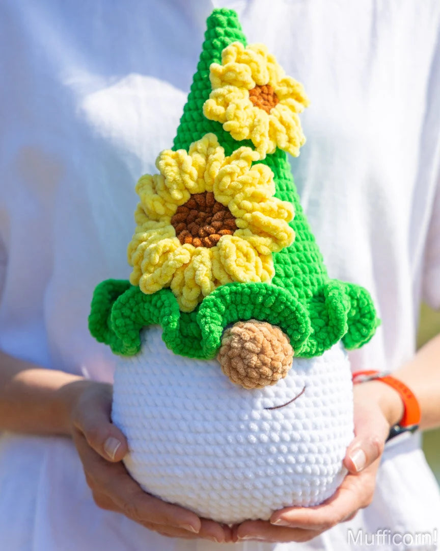 A crocheted sunflower gnome doll being held in both hands. The doll has a white textured body, a green pointed hat decorated with two yellow sunflowers, and a brown yarn nose.