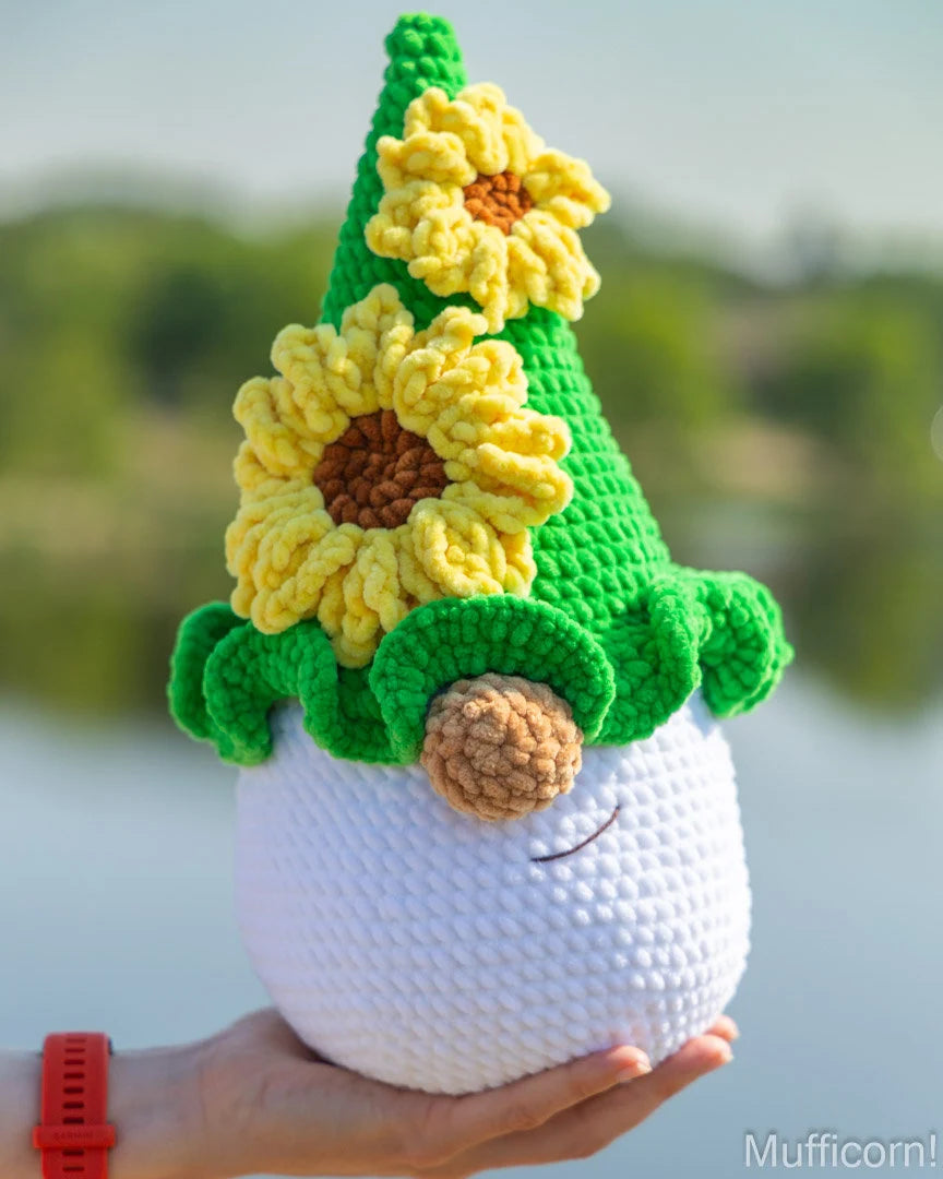 A crocheted sunflower gnome doll being held in a person's hand outdoors. The doll has a white body, a green pointed hat with yellow sunflowers, and a brown nose.