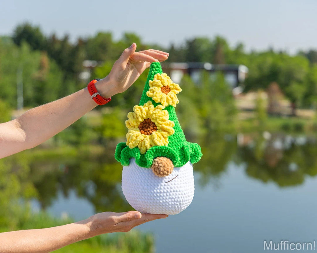 A crocheted plush gnome with yellow sunflowers and a green pointed hat, held up by two hands outdoors in front of a blurred lake and trees. The gnome has a white body, brown nose, and black embroidered eyes.
