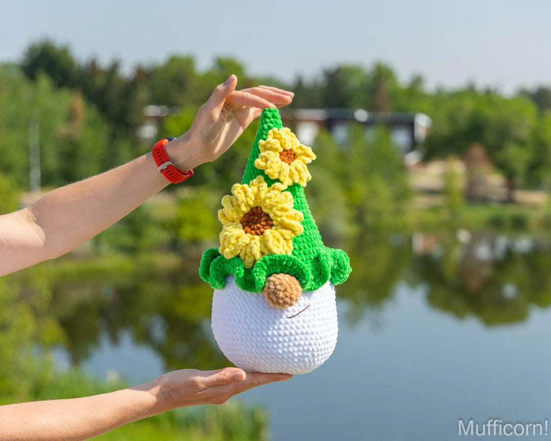 A crocheted plush gnome with yellow sunflowers and a green pointed hat, held up by two hands outdoors in front of a blurred lake and trees. The gnome has a white body, brown nose, and black embroidered eyes.