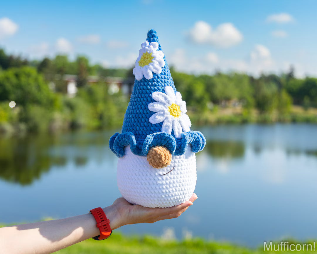 A crocheted gnome doll with a blue hat, white body, and daisy details is held in a person's hand outdoors in front of a lake and trees.