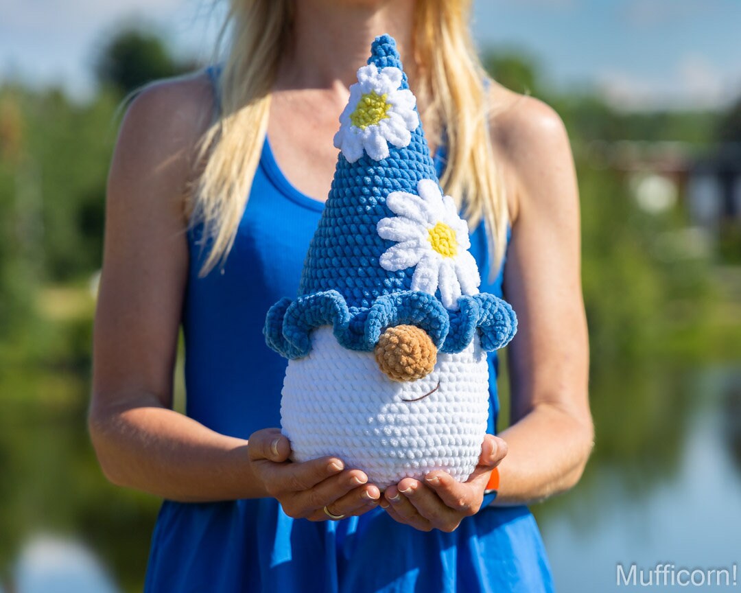 A person in a blue dress holds a crocheted plush gnome doll outdoors with a blurred lake and trees in the background. The gnome has a blue pointed hat with two white daisy flowers with yellow centers, a blue ruffled collar, a white body, and a brown carrot nose.