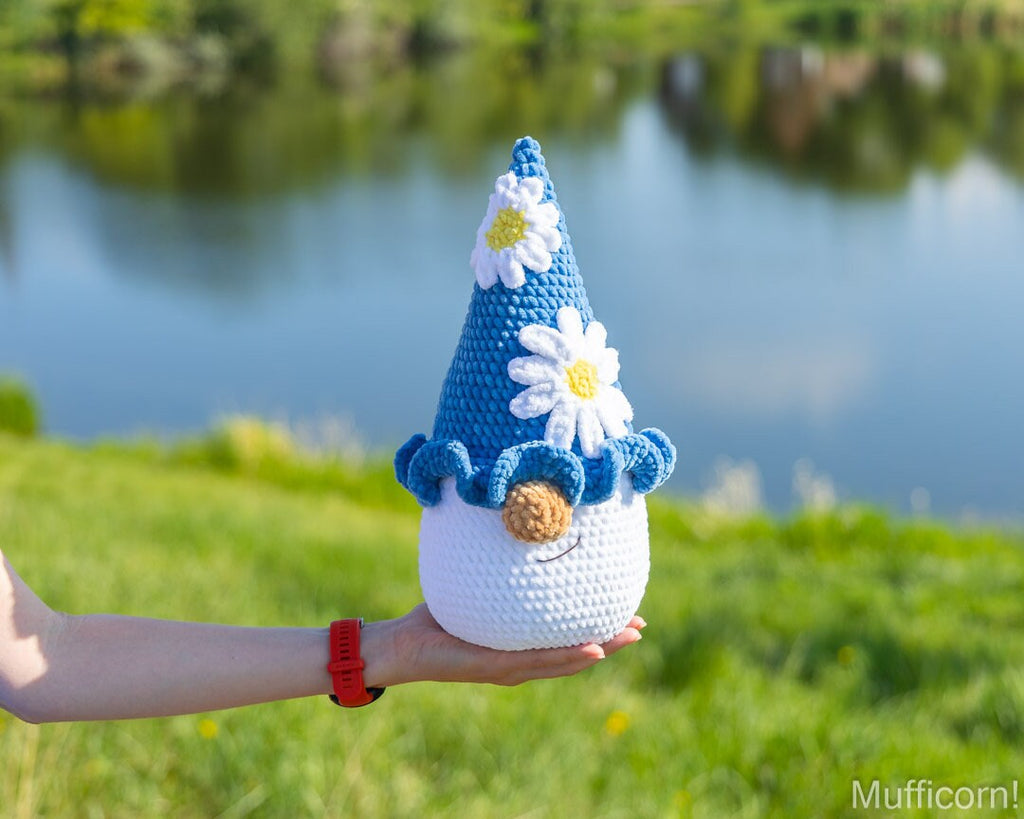 A crocheted plush gnome doll with a blue pointed hat decorated with white daisies, held in a person's hand against a blurred outdoor background of green grass and a blue lake.