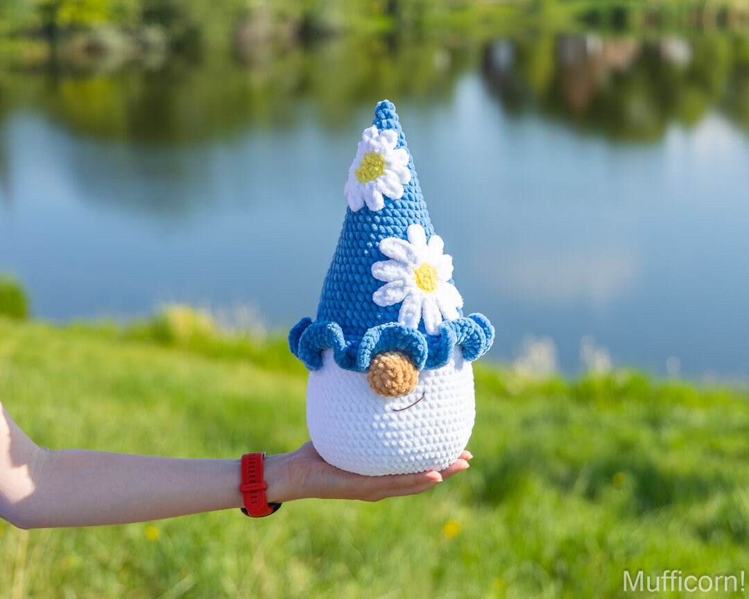 A crocheted plush gnome doll with a blue pointed hat decorated with white daisies, held in a person's hand against a blurred outdoor background of green grass and a blue lake.