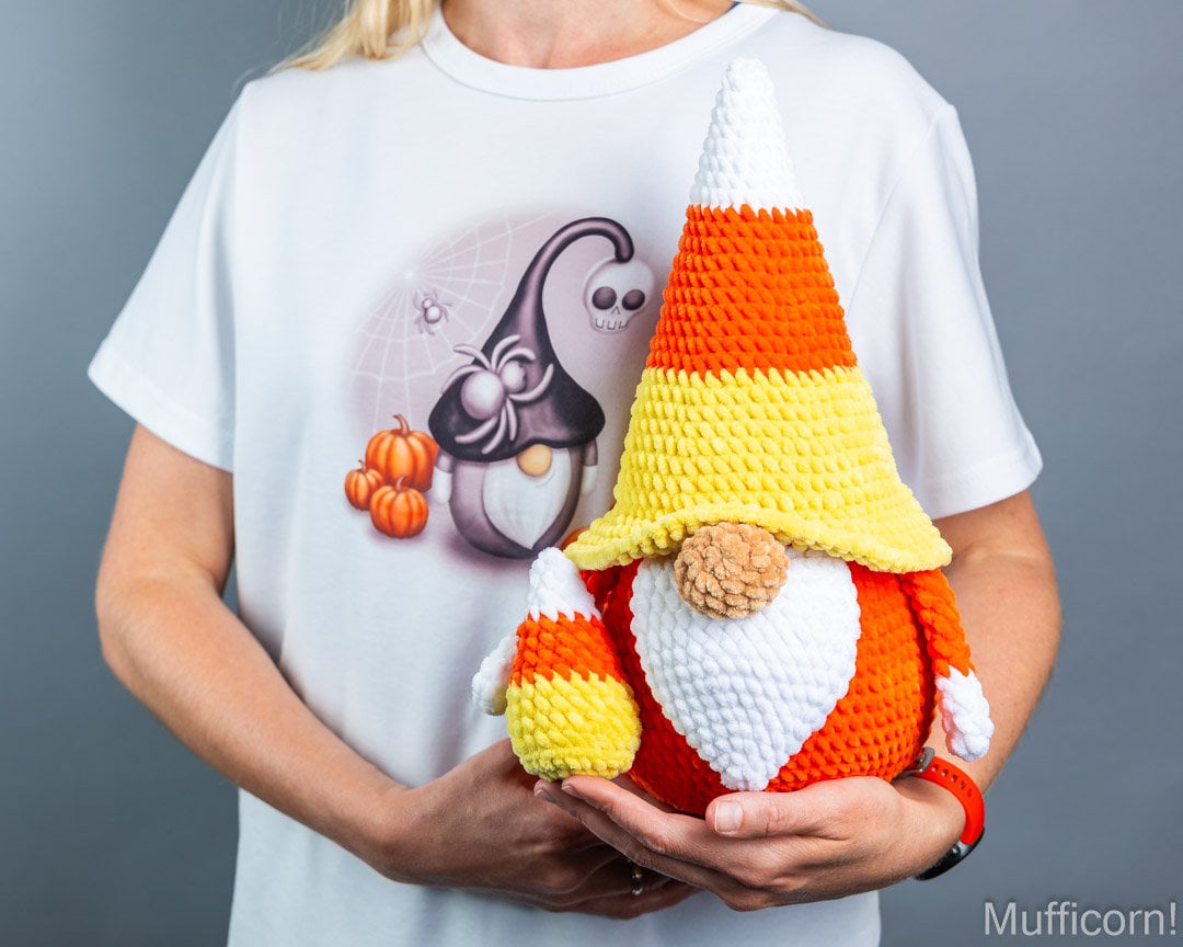 A person wearing a white graphic T-shirt holds a crocheted candy corn gnome. The gnome has a pointed hat and beard made of orange, yellow, white, and brown yarn, with a small matching crocheted candy corn accessory in one hand.