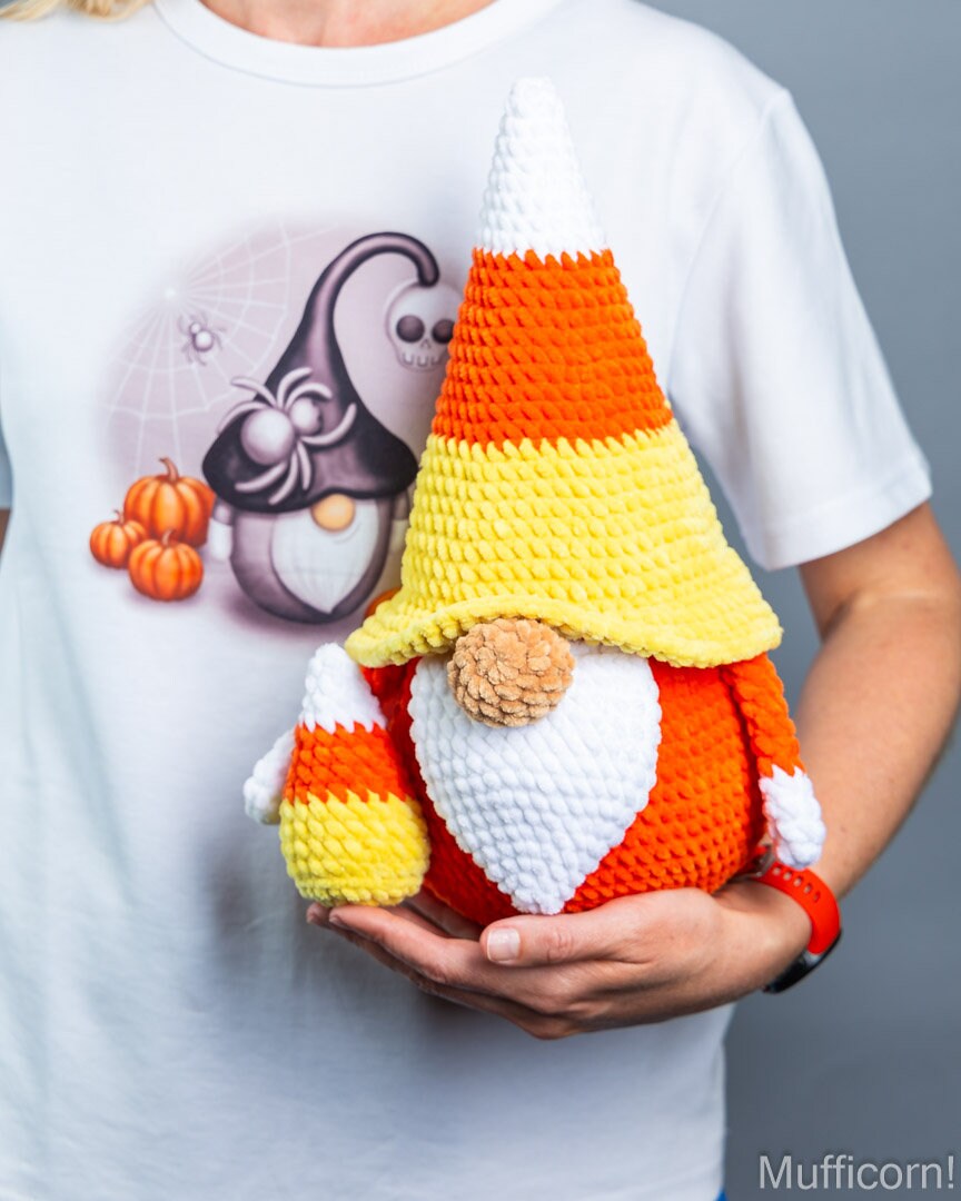 A person in a white graphic t-shirt holds a crocheted candy corn gnome doll. The gnome has a pointed hat and beard made of orange, yellow, and white yarn, with a small pinecone for a nose.