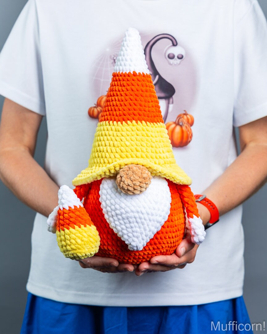 A person wearing a white graphic t-shirt holds a crocheted candy corn gnome. The gnome features a pointed hat striped in orange, yellow, and white, a white beard, and an orange body, with a small brown yarn nose.