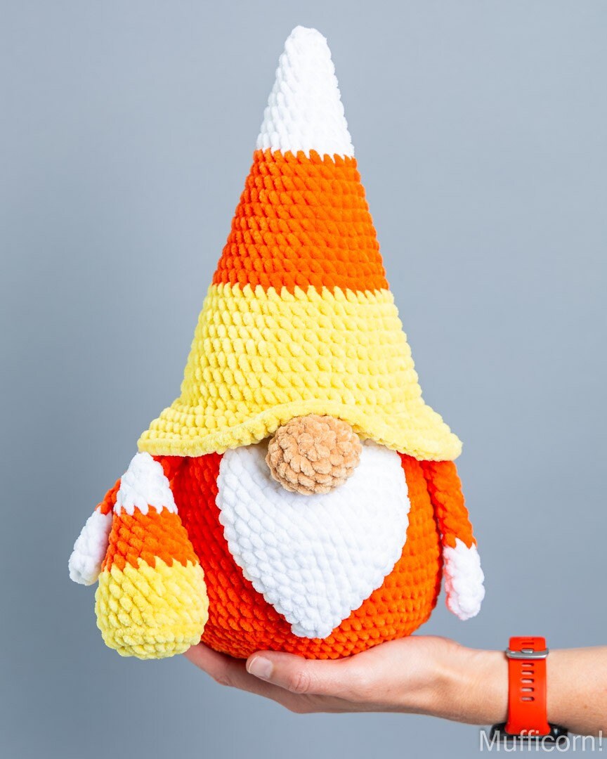A crocheted plush candy corn gnome being held in a person's hand against a plain gray studio background. The gnome has a pointed hat and beard in white, yellow, and orange, with small striped arms and a tan nose.