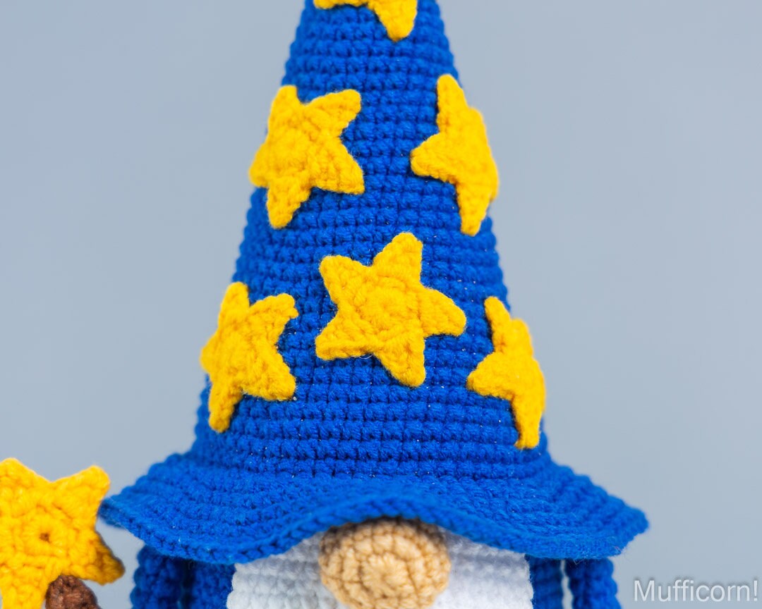 Close-up of a crocheted wizard gnomehat in bright blue with yellow star appliqués, shown against a soft light gray background.