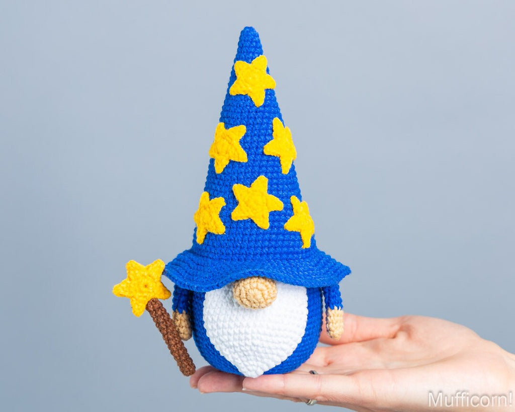 A crocheted wizard gnome doll with a tall blue hat decorated with yellow stars, held in a person's hand against a plain light gray background.