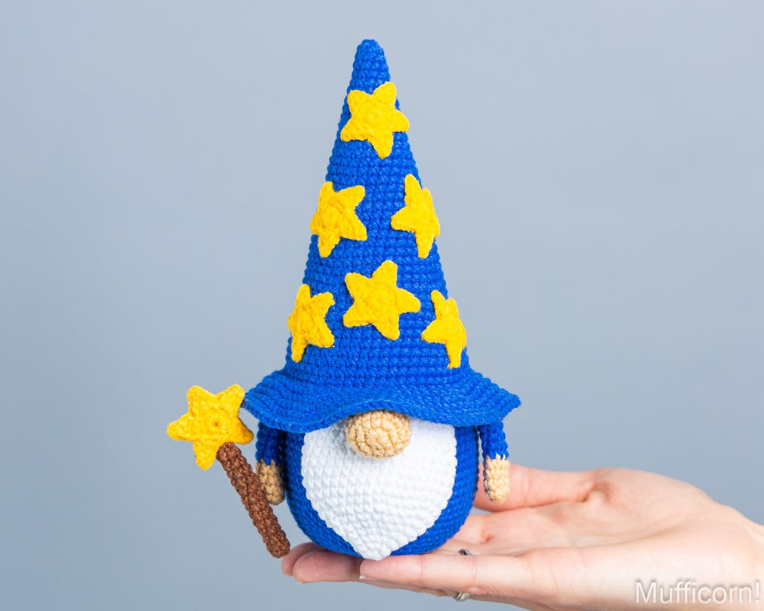 A crocheted wizard gnome doll with a tall blue hat decorated with yellow stars, held in a person's hand against a plain light gray background.