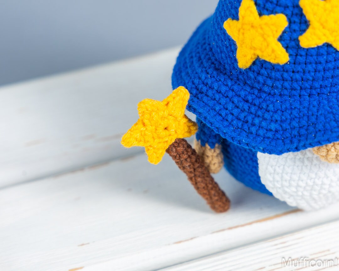 Close-up of a crocheted wizard character wearing a blue hat with yellow stars and holding a brown staff topped with a yellow star, set on a white wooden surface.