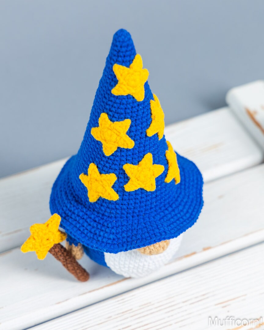 A crocheted wizard gnome figurine with a tall blue hat decorated with yellow felt stars, a white beard, and a brown staff, displayed on a white wooden surface against a soft gray background.