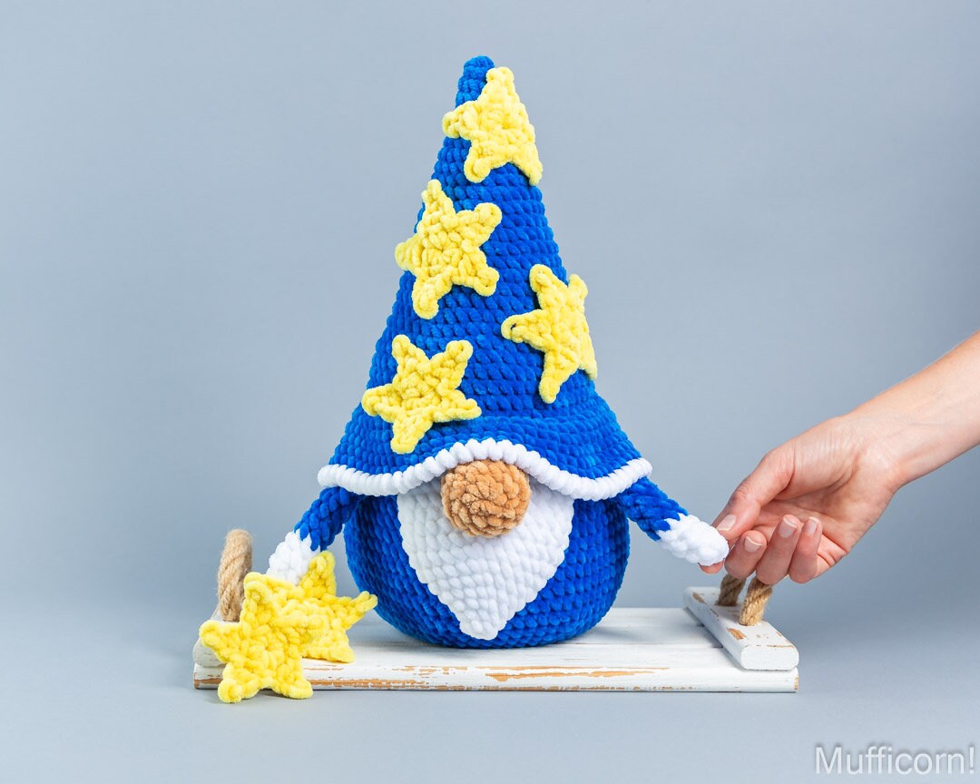 A crocheted gnome figure with a tall blue hat decorated with yellow stars, a white beard, and a brown nose, posed on a small white platform. A human hand is holding one of the star ornaments beside the gnome against a plain light gray studio background.