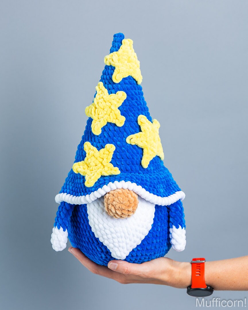 A handmade crocheted gnome doll with a tall blue hat decorated with yellow stars, held in a person's hand against a solid gray background.