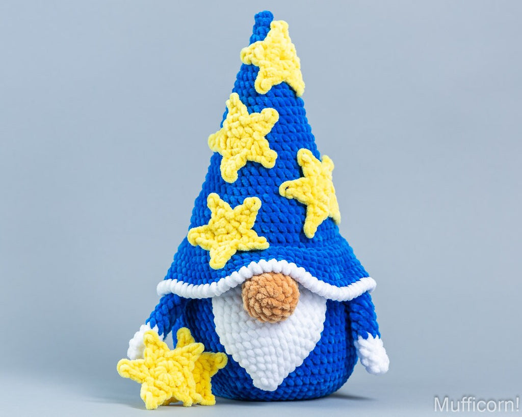 A crocheted gnome figurine with a tall blue hat decorated with yellow stars, a white beard, and a brown nose, photographed against a plain light gray background.