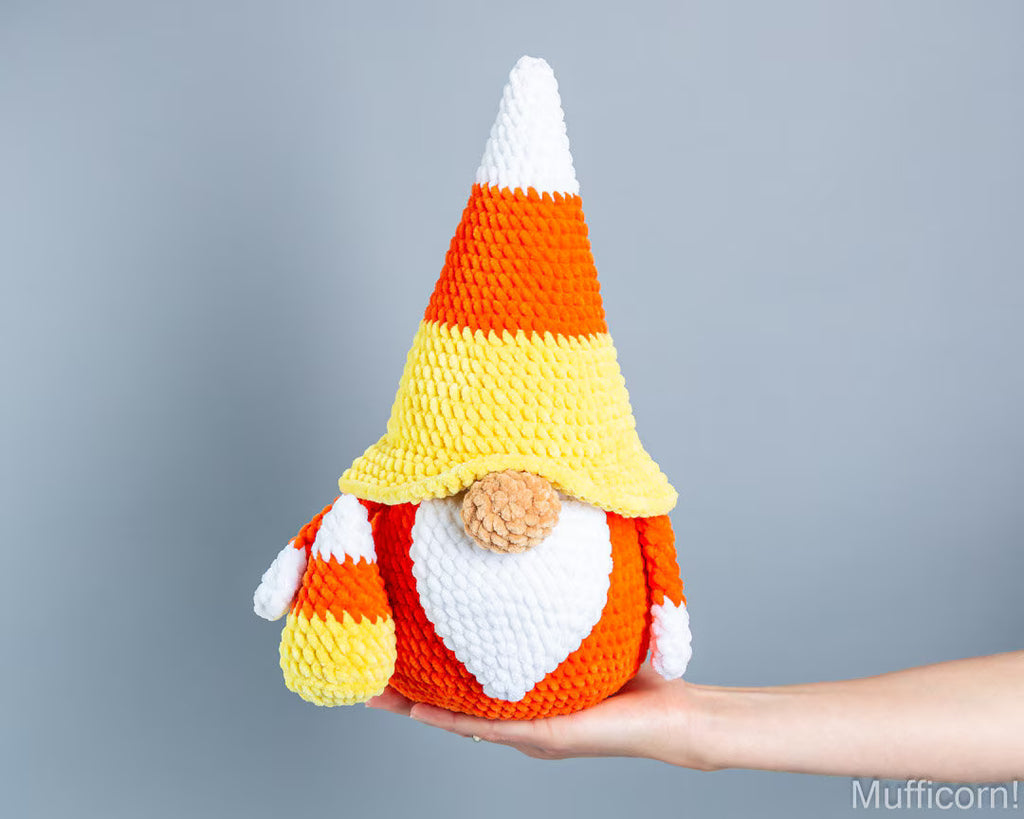 A crocheted candy corn gnome doll with a pointed hat and beard, held in a person's hand against a plain gray background.