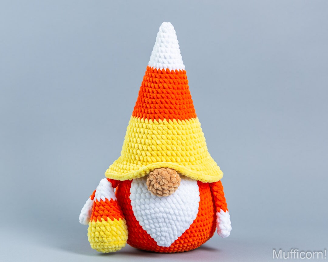 A crocheted candy corn gnome doll with a pointed hat and matching small bag, shown against a plain light gray studio background.