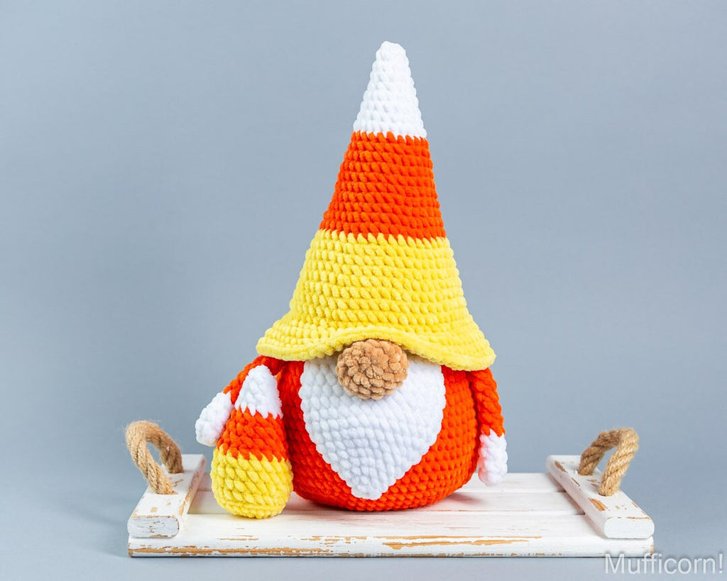 A crocheted candy corn gnome sits on a white wooden sled against a solid gray background. The gnome is designed like a candy corn cone with white, yellow, and orange stripes, a white beard, and a small brown pom-pom nose.