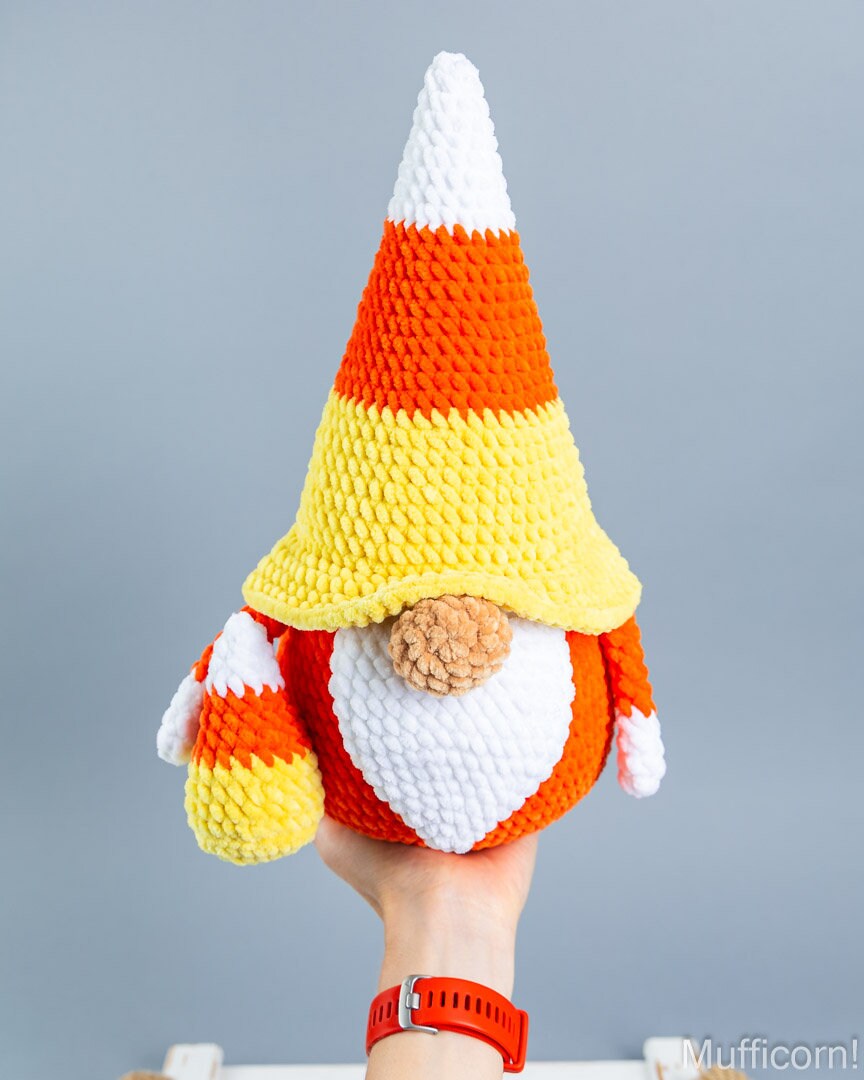 A crocheted candy corn gnome doll held up by a person's hand against a plain gray background. The gnome has a pointed hat and sleeves in white, orange, and yellow, with a white beard and a brown nose.