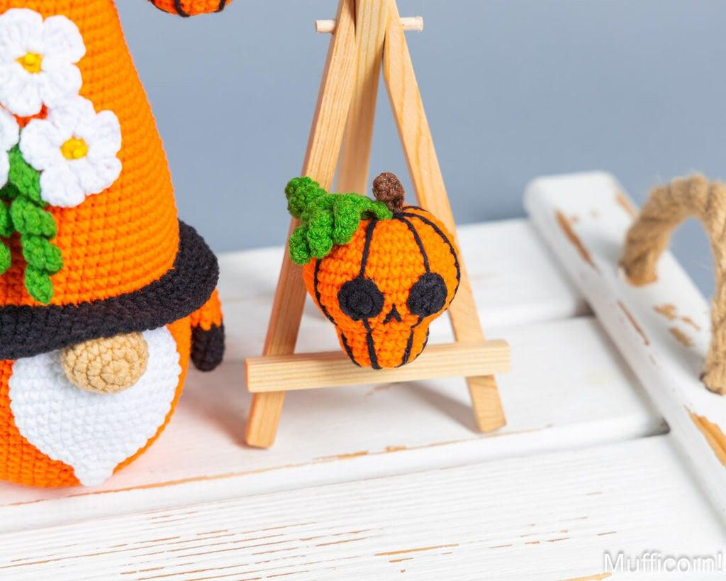 A crocheted orange pumpkin skull with a black stitched face and green leaves, displayed on a small wooden easel. Part of an orange gnome figure with white flowers is visible on the left, and a rustic white wooden prop sits on the right against a soft blue-gray background.