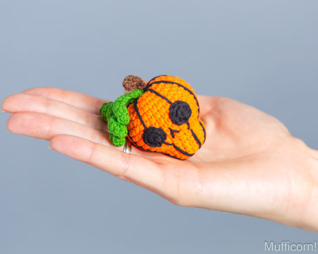 A small crocheted jack-o'-lantern keychain with a green stem rests on an open hand against a solid gray background.