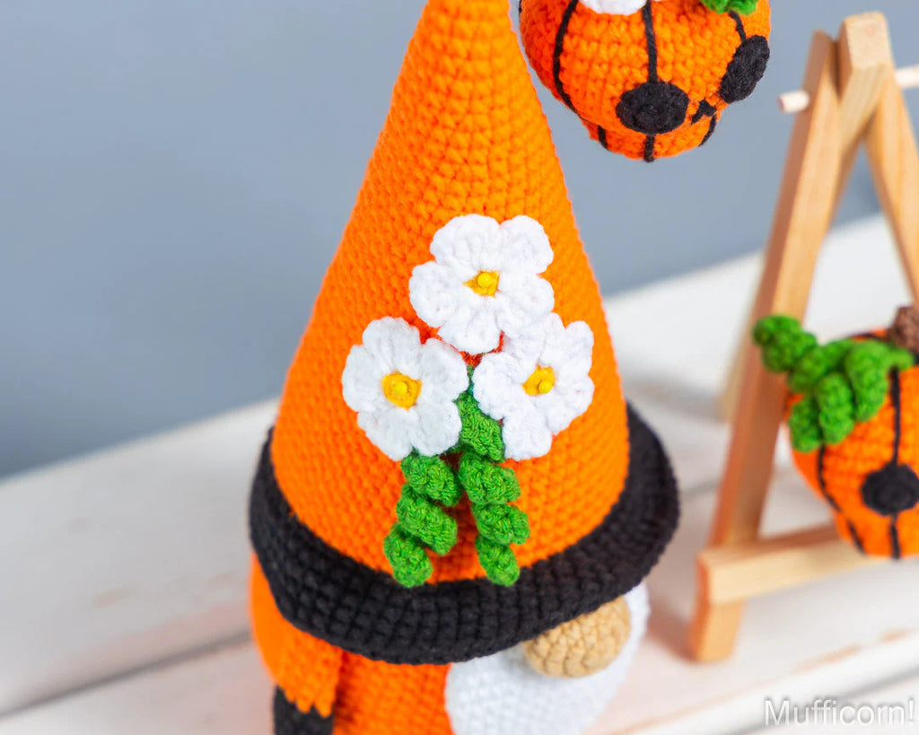 A crocheted orange gnome hat with white daisy flowers, green stems, and a black band, shown close up against a soft blue-gray background.