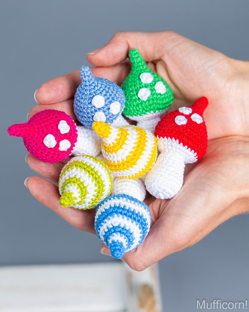 A pair of hands holding a cluster of small crocheted mushroom-shaped ornaments in multiple bright colors, including pink, blue, green, yellow, red, and white, against a soft gray background.