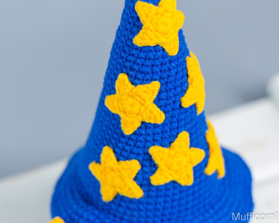 A close-up of a blue crocheted wizard hat with yellow star appliqués, photographed against a soft light gray background.