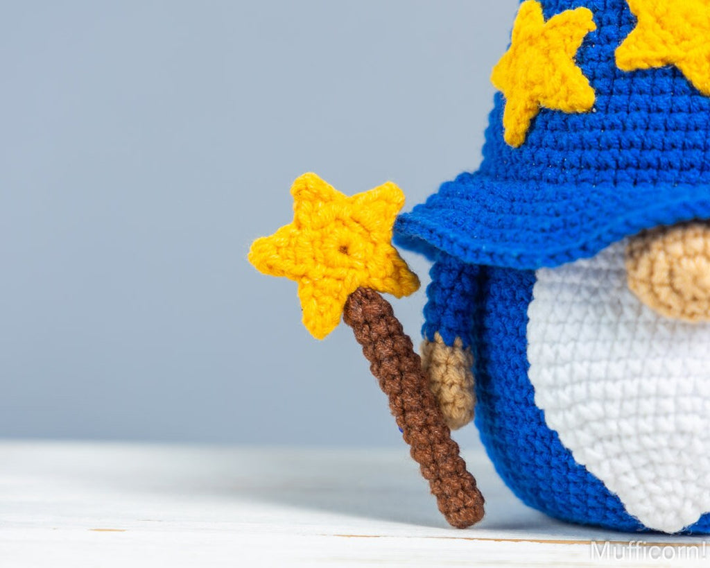 A crocheted wizard gnomemall with a blue hat and cape, white beard, and yellow stars, holding a brown and yellow star wand, shown against a light gray background.