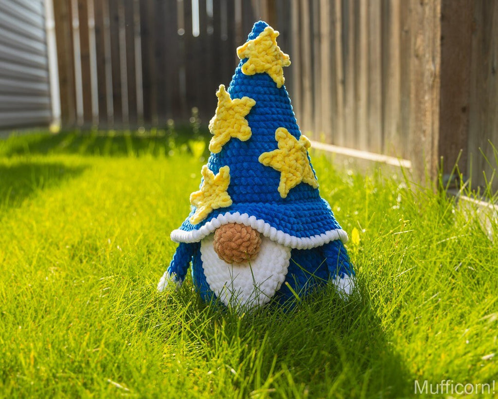 A crocheted gnome wearing a tall blue hat with yellow stars, standing in bright green grass outdoors near a wooden fence. The gnome has a white beard, brown nose, and blue shoes.