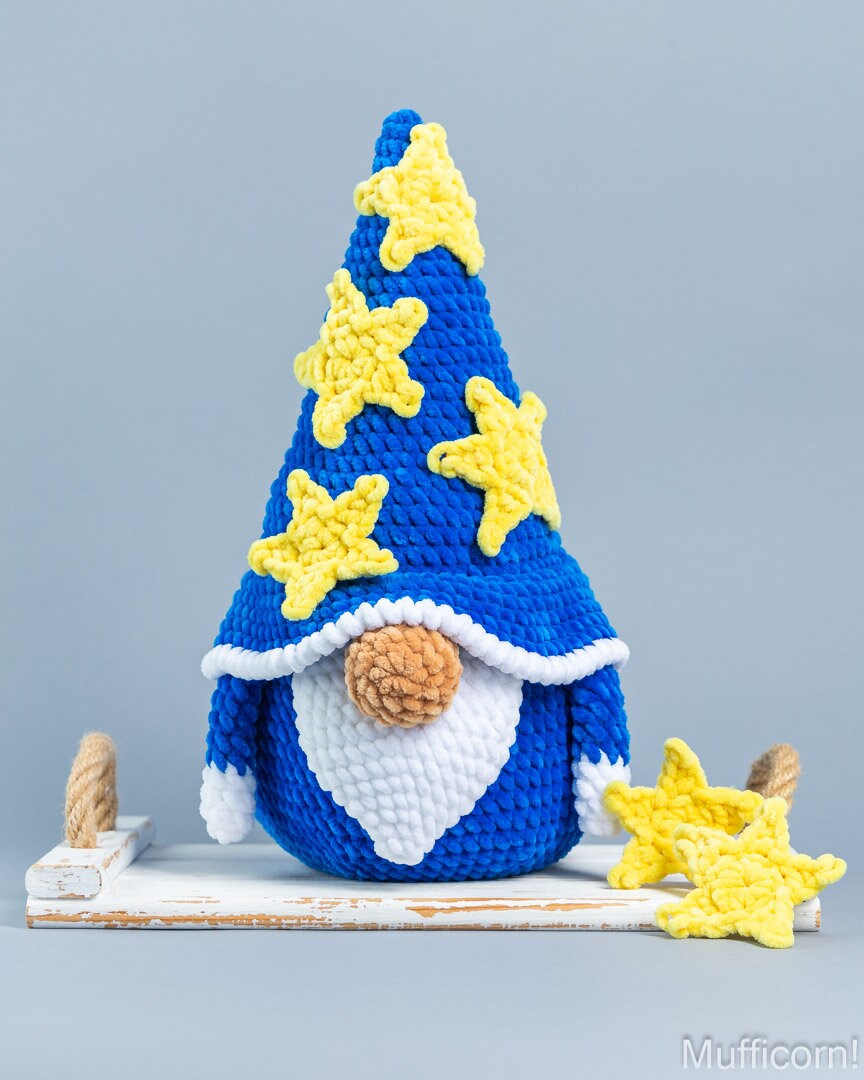 A crocheted gnome figurine with a tall blue hat decorated with yellow stars, a white beard, and a brown pom-pom nose, standing on a small white platform against a light gray background.