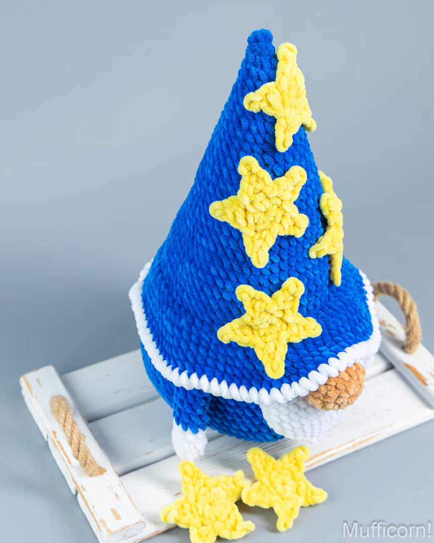 A blue knit gnome hat with yellow crocheted stars and a white trim, displayed on a white wooden crate against a gray background.